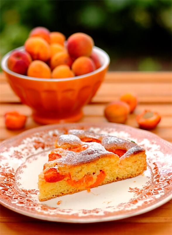 Aprikoosikakku pala lautaselle sen vieressä tuoreita aprikooseja hedelmäkulhoon