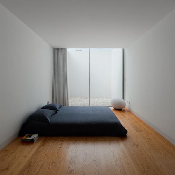 Kalusta makuuhuone minimalistisella tavalla. Suuri kattoon ulottuva ikkuna. Harmaat seinät. Musta sänky. Lattia vaaleaa puuta