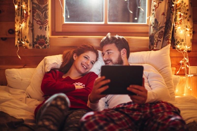 Pari makaa viihtyisällä sängyllä puumökissä. Joulupyjama päällä. Yhdessä katsomassa elokuvaa digitaalisella tabletilla. Nauttivat kodin lämmöstä jouluna. Huone on sisustettu juhlavaloilla. Itävallan Alpit. Illalla tai yöllä kauniit keltaiset valot salamoivat kohtauksia.